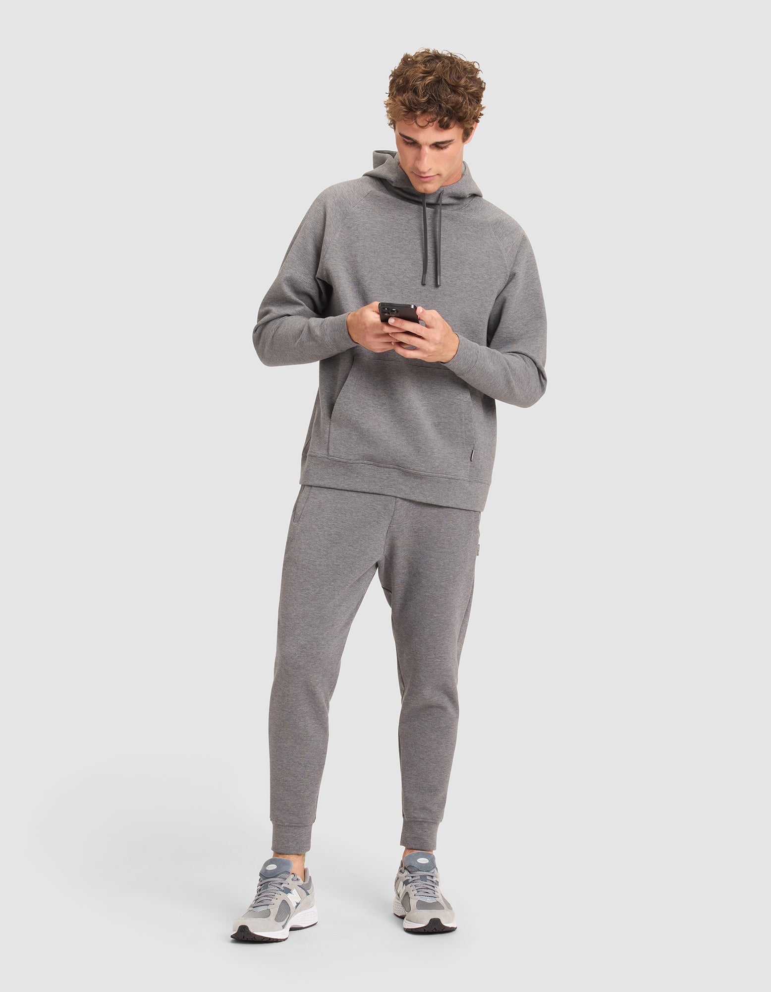 A man with curly hair models the Cozy Earth Men's StretchTech Hoodie & Jogger Set in gray, paired with white and gray sneakers, as he looks down at his smartphone against a plain white background.  