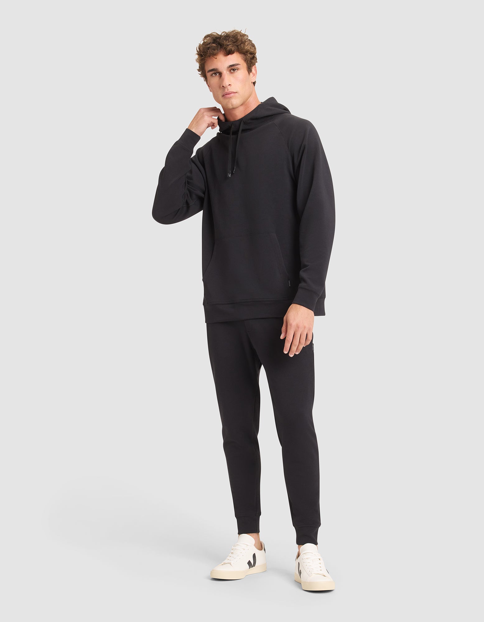 A young man with short curly hair poses against a light gray background, wearing the Cozy Earth Men's StretchTech Hoodie & Jogger Set in black with white sneakers, looking at the camera and touching his neck. |Color:Jet Black