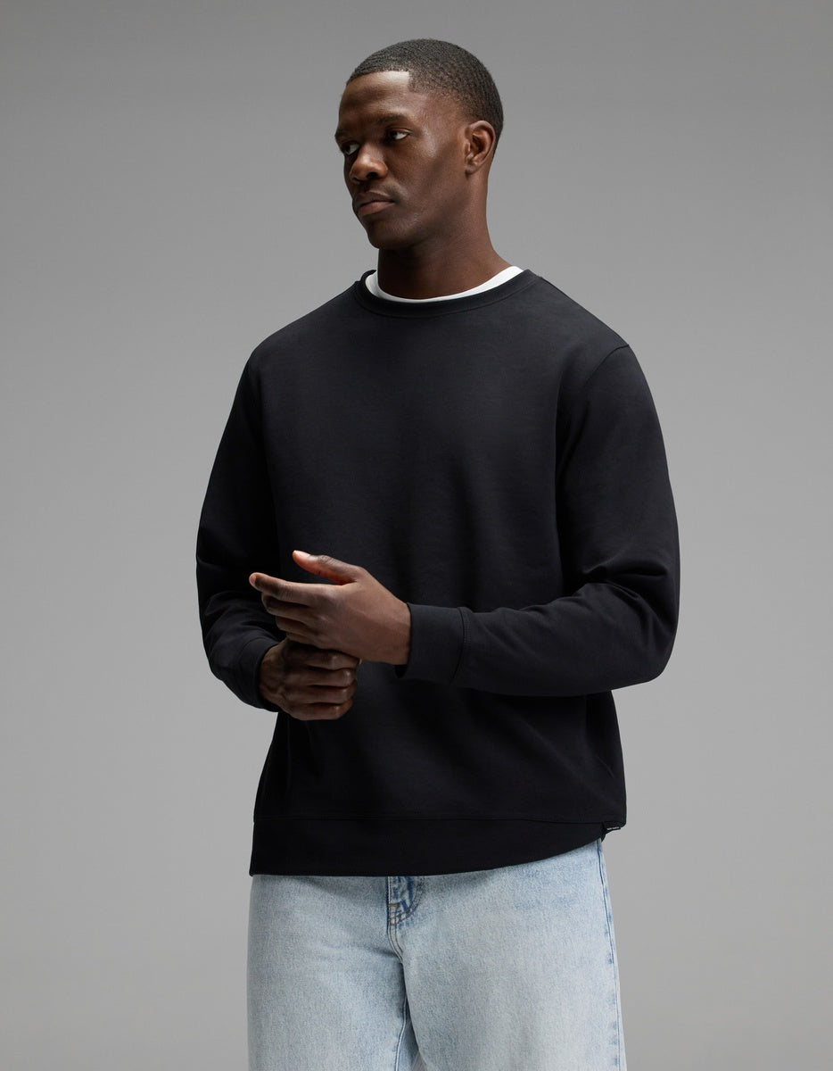 A person wearing the Cozy Earth Men’s StretchTech Crewneck in Jet Black over a white shirt and light blue jeans stands against a plain gray background, looking slightly to the side with hands together.|Color:Jet Black