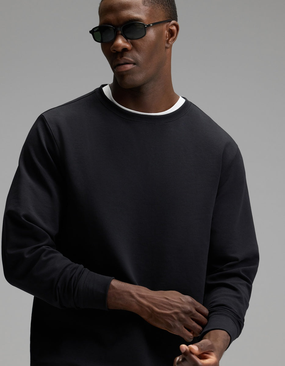 A man wearing dark sunglasses and a black Cozy Earth Men’s StretchTech Crewneck over a white shirt stands against a plain gray background.