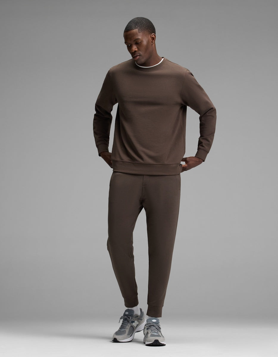 A man stands against a plain gray background, wearing the Cozy Earth Men’s StretchTech Crewneck & Jogger Set in brown, paired with gray running shoes. He looks down, hands in his pockets.|Color:Cacao