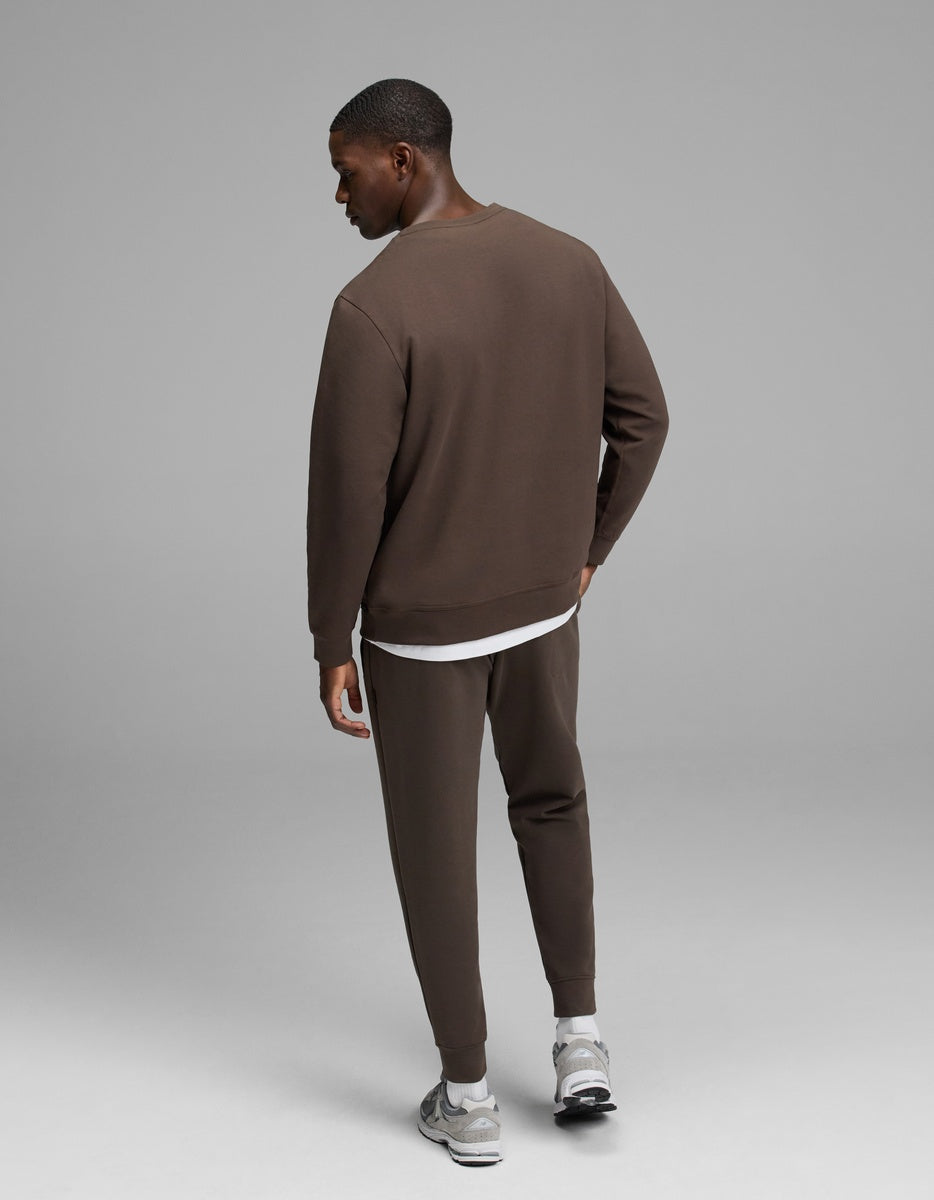 A man stands facing away, wearing Cozy Earth’s Men’s StretchTech Crewneck & Jogger Set in brown over a white shirt, paired with gray sneakers, against a plain gray background.