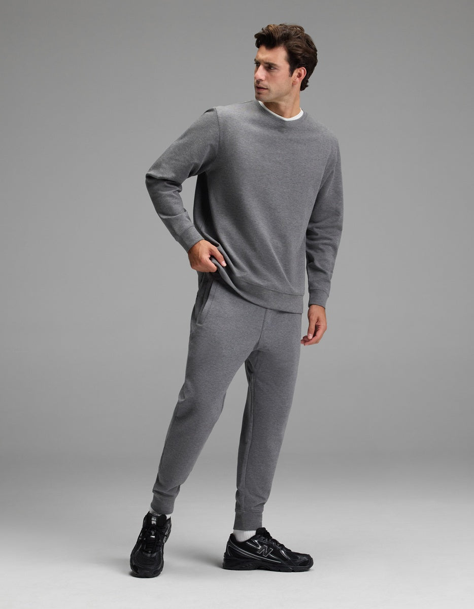 A man wearing the Cozy Earth Men’s StretchTech Crewneck & Jogger Set and black sneakers stands against a plain grey background, looking to his right with one hand in his pocket.