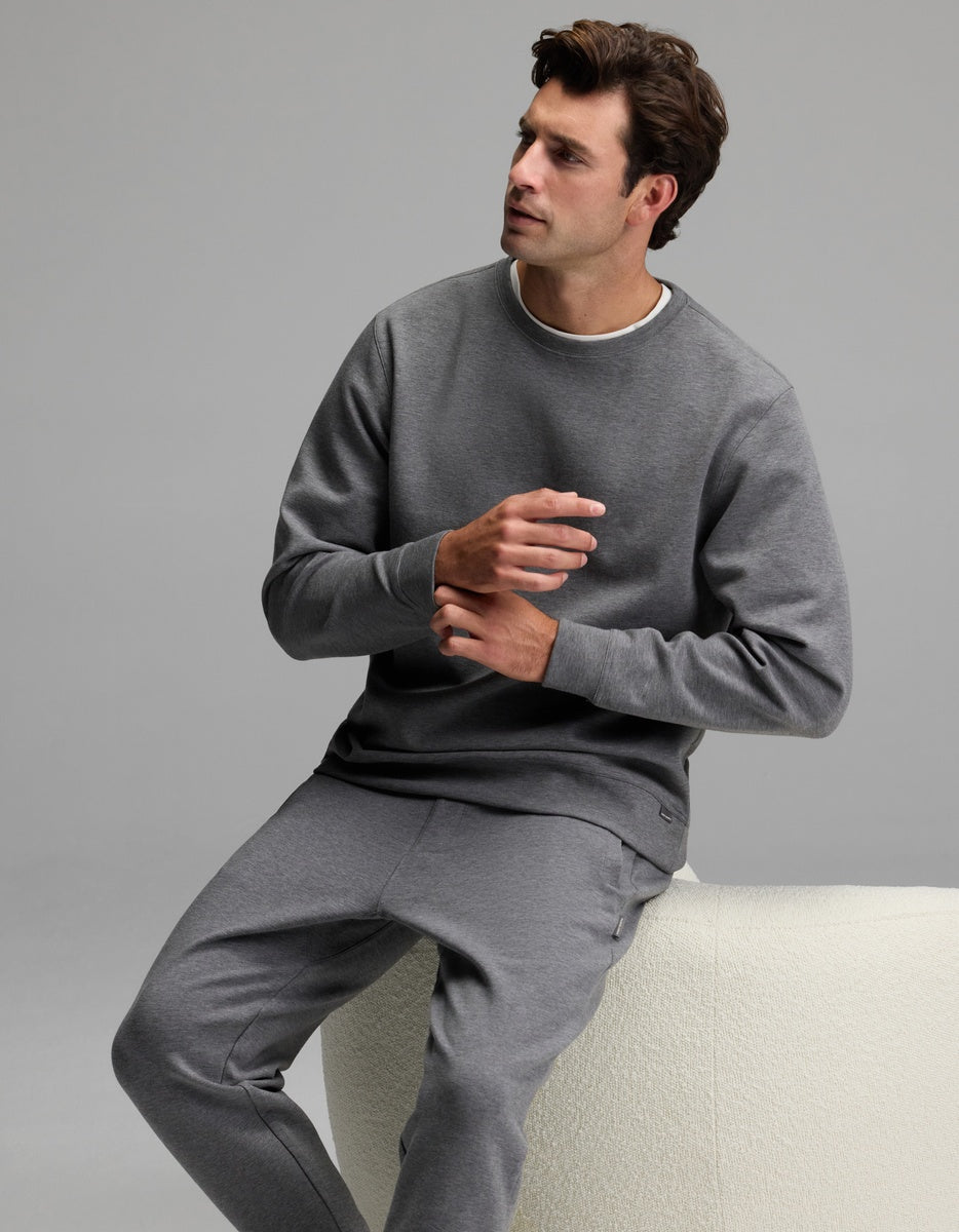 A man wears the Cozy Earth Men’s StretchTech Crewneck & Jogger Set in gray as he sits on a light-colored bench, looking to his left with his hands gesturing in front of him against a plain, neutral background.