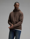 A man wearing the Cozy Earth Men’s StretchTech Hoodie and blue jeans stands against a plain gray background, looking to his left with his hands loosely clasped in front of him.|Color:Cacao