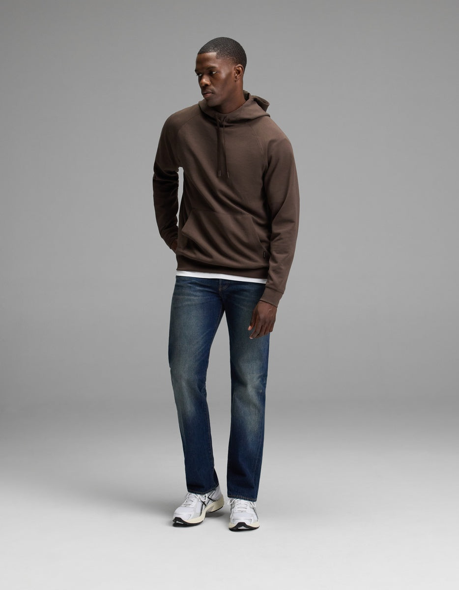 A person stands in a studio wearing the Cozy Earth Men’s StretchTech Hoodie, paired with blue jeans and white sneakers. They look down and to the side with their hands relaxed by their sides against a plain gray background.