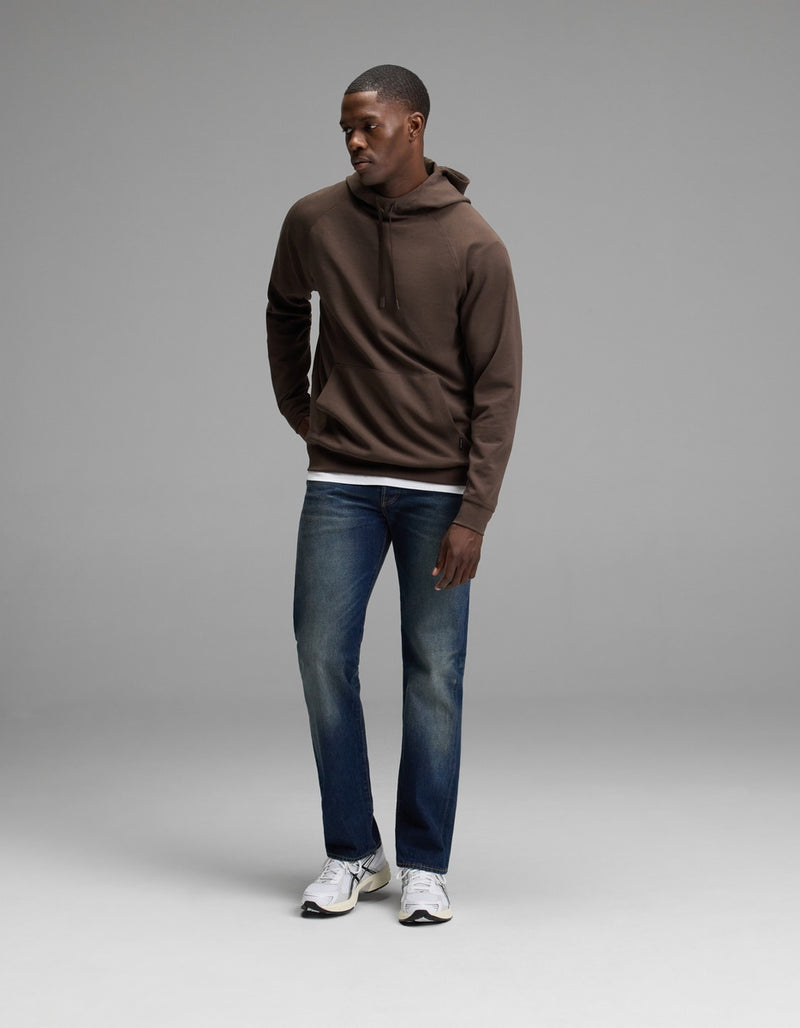 A person stands in a studio wearing the Cozy Earth Men’s StretchTech Hoodie, paired with blue jeans and white sneakers. They look down and to the side with their hands relaxed by their sides against a plain gray background.