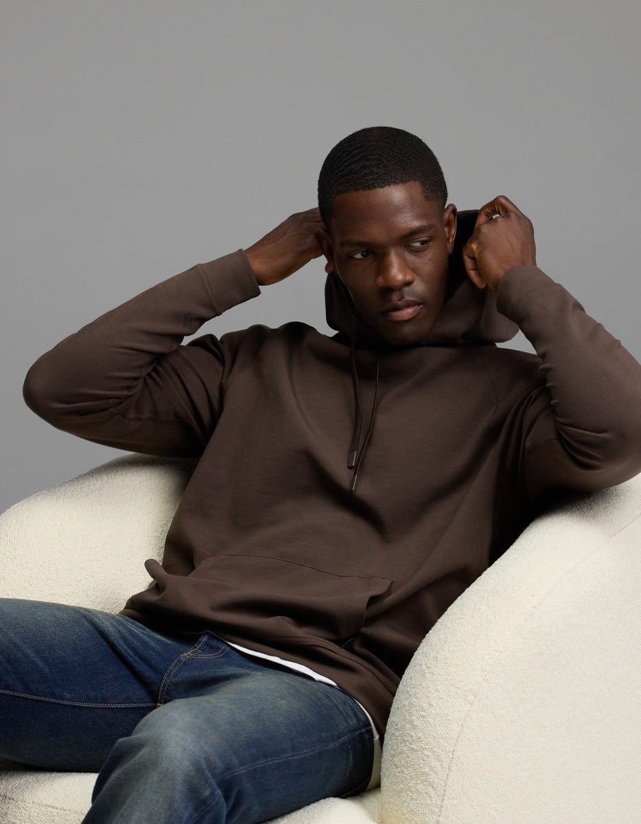 A man wearing the Cozy Earth Men’s StretchTech Hoodie in brown and blue jeans sits on a white chair, adjusting his hood with both hands while looking slightly to the side against a plain gray background.