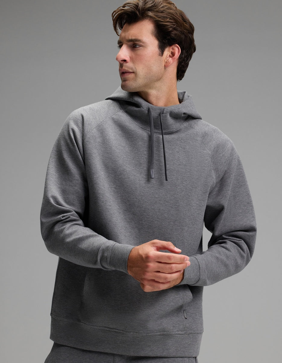 A man wearing the Cozy Earth Men’s StretchTech Hoodie in gray stands against a plain gray background, looking to his left with his hands clasped.