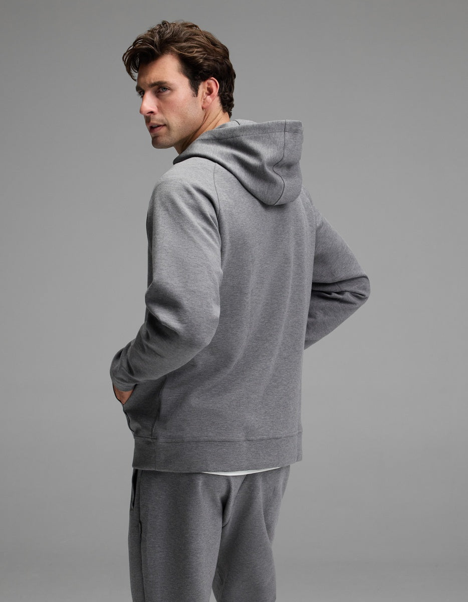 A man wearing the Cozy Earth Men’s StretchTech Hoodie and matching sweatpants stands with his back mostly to the camera, turning his head to look over his left shoulder against a plain, light gray background.