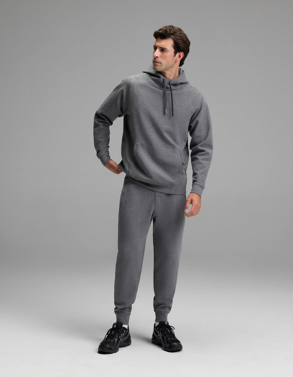 A man stands in a studio wearing the Cozy Earth Men’s StretchTech Hoodie in gray, paired with matching sweatpants and black athletic shoes. He looks to his left with one hand on his hip against a plain, light gray background.
