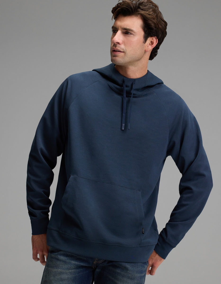 A man wearing the Cozy Earth Men’s StretchTech Hoodie in navy blue and jeans stands with one hand in his pocket against a plain gray background.