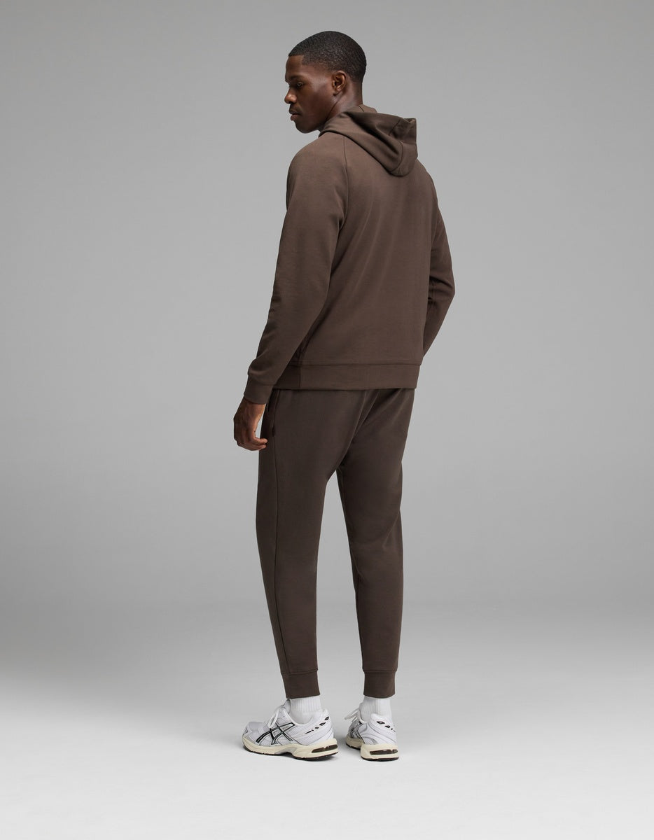 A man stands facing away, wearing the Cozy Earth Men’s StretchTech Hoodie & Jogger Set in brown with white sneakers, against a plain gray background.