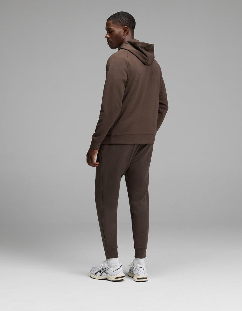 A man stands facing away, wearing the Cozy Earth Men’s StretchTech Hoodie & Jogger Set in brown with white sneakers, against a plain gray background.