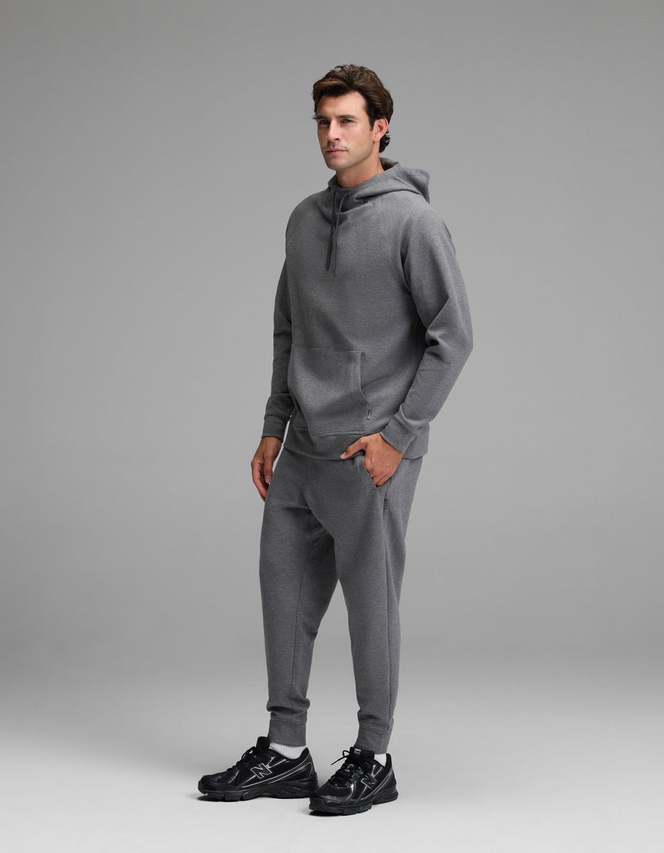 A man stands in a studio wearing the Cozy Earth Men’s StretchTech Hoodie & Jogger Set in gray, paired with black sneakers. The background is plain and light gray.