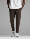 A person is shown from the waist down wearing Cozy Earth’s Men’s StretchTech Jogger in dark brown, paired with a white T-shirt and white sneakers, standing against a plain gray background.|Color:Cacao