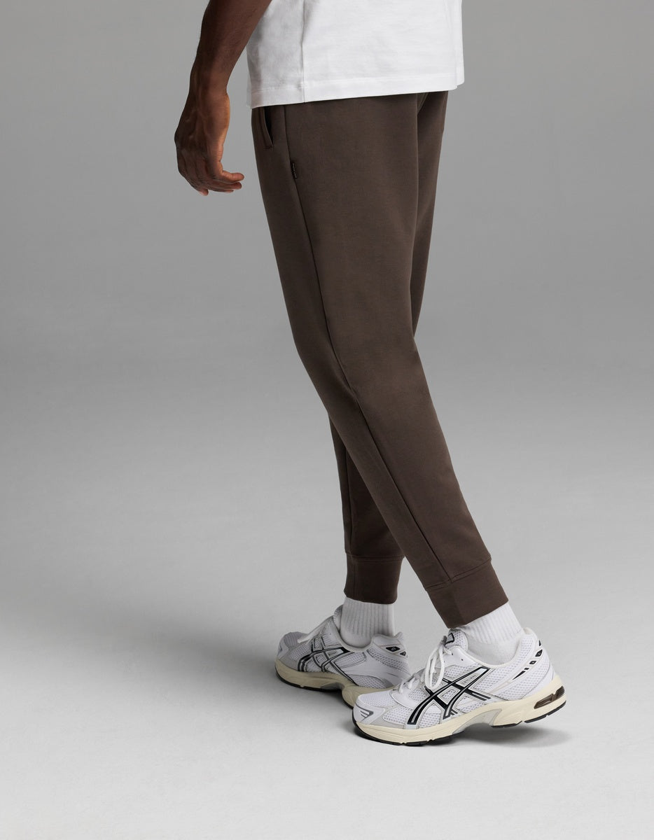 A person is shown from the waist down, standing against a plain gray background, wearing Cozy Earth’s Men’s StretchTech Jogger in brown, paired with a white T-shirt, white socks, and white sneakers.