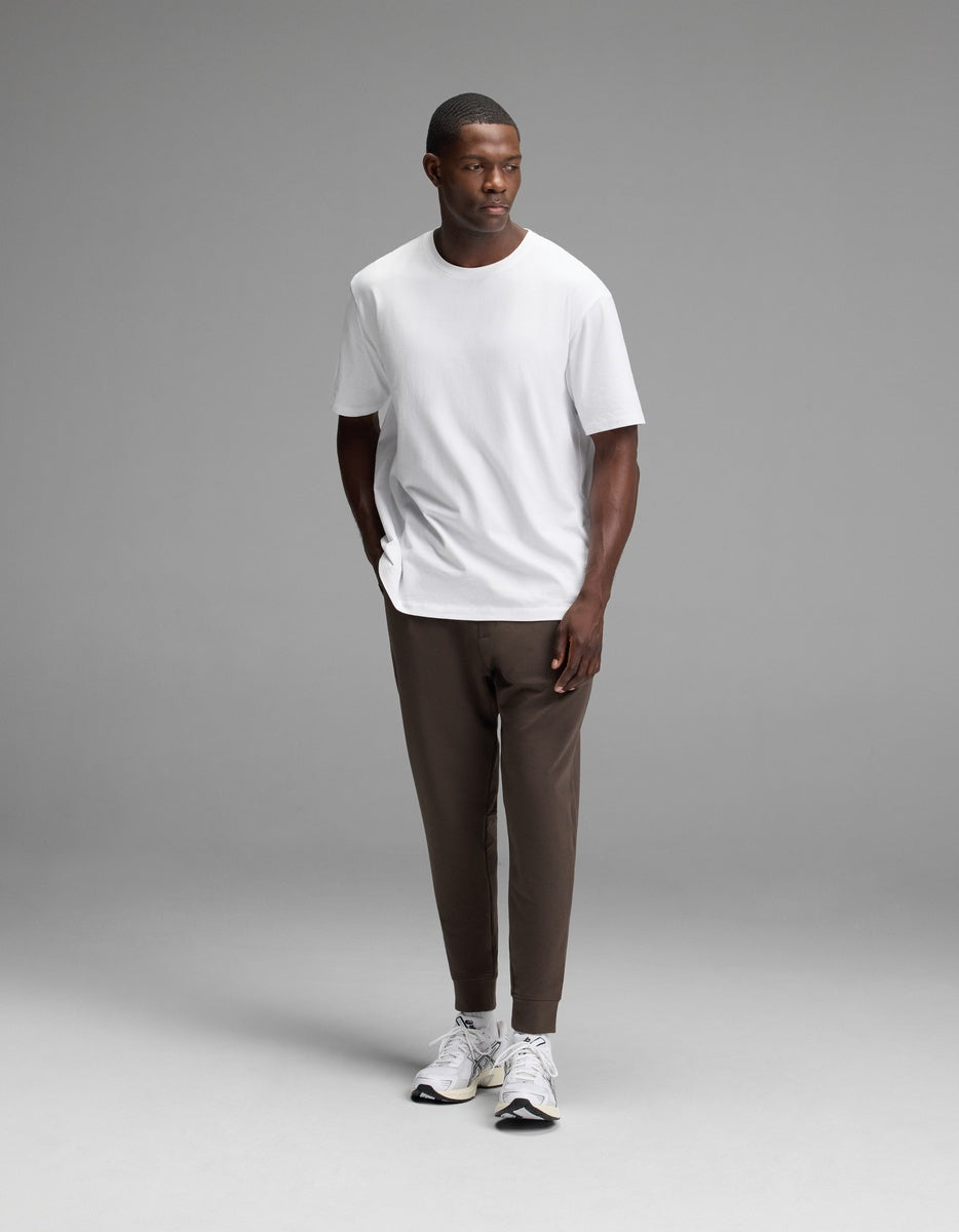 A man stands against a plain gray background wearing a white T-shirt, Cozy Earth Men’s StretchTech Jogger in brown, and white sneakers, with his hands in his pockets.