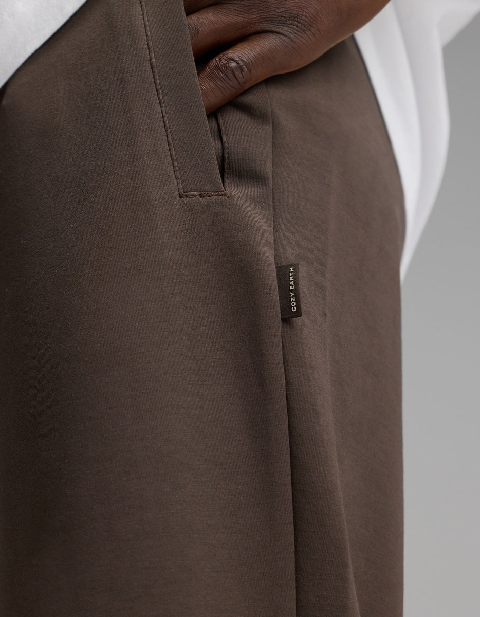 A close-up of a person’s hand in the pocket of Cozy Earth Men’s StretchTech Jogger pants in brown, featuring a small fabric tag with text. The person is also wearing a white shirt.