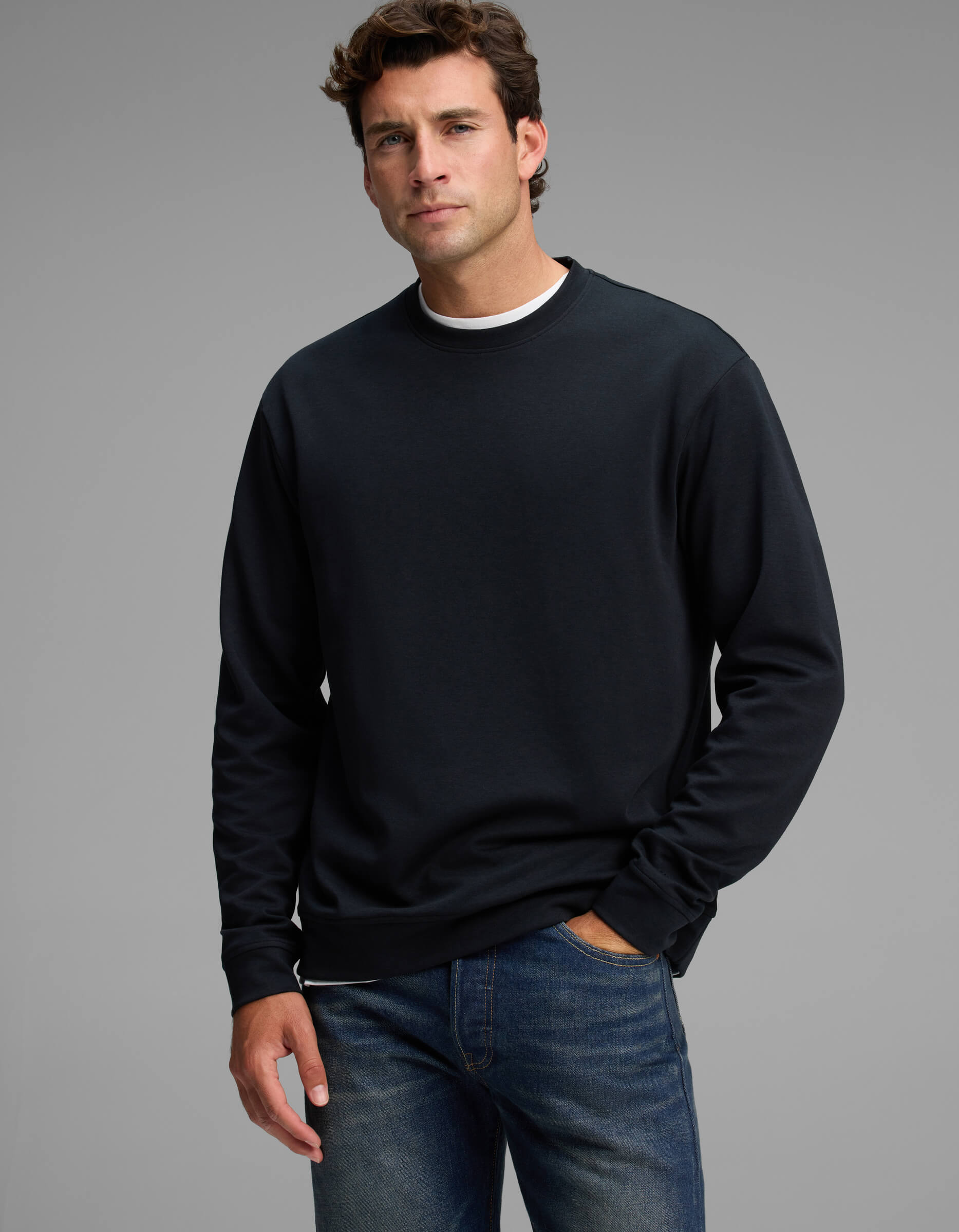 A man stands against a gray background, wearing the Cozy Earth Men's Versa Crewneck in black over a white shirt and blue jeans, with one hand in his pocket and a neutral expression.