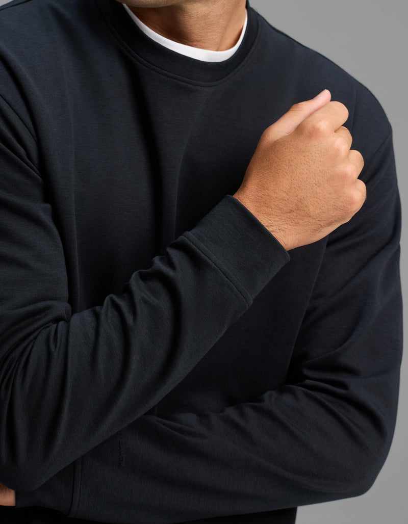 A person is shown from the shoulders down wearing the Cozy Earth Men's Versa Crewneck in black, with one arm crossed and the other bent, hand near the chest, against a gray background.