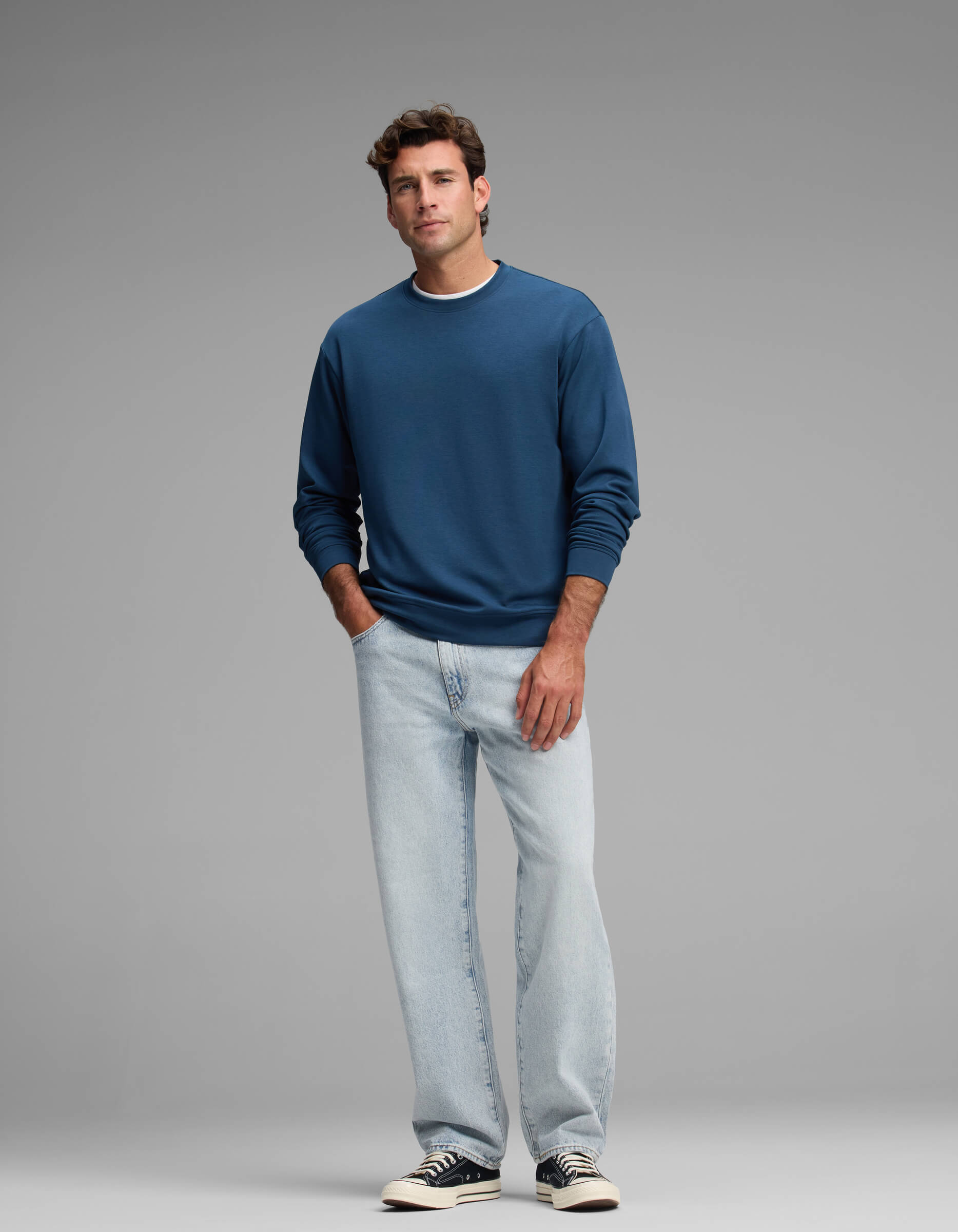 A man stands against a plain gray background wearing the Cozy Earth Men's Versa Crewneck, light blue jeans, and black-and-white sneakers.