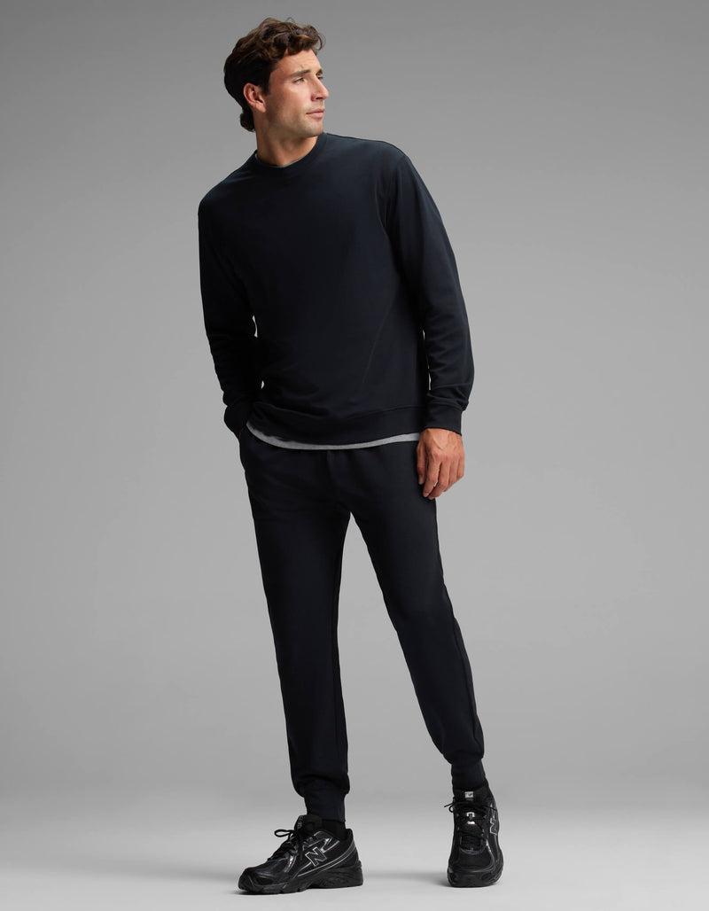 A man stands in a relaxed pose against a plain gray background, wearing the Cozy Earth Men's Versa Crewneck & Jogger Set in black, paired with black sneakers and a visible gray undershirt.