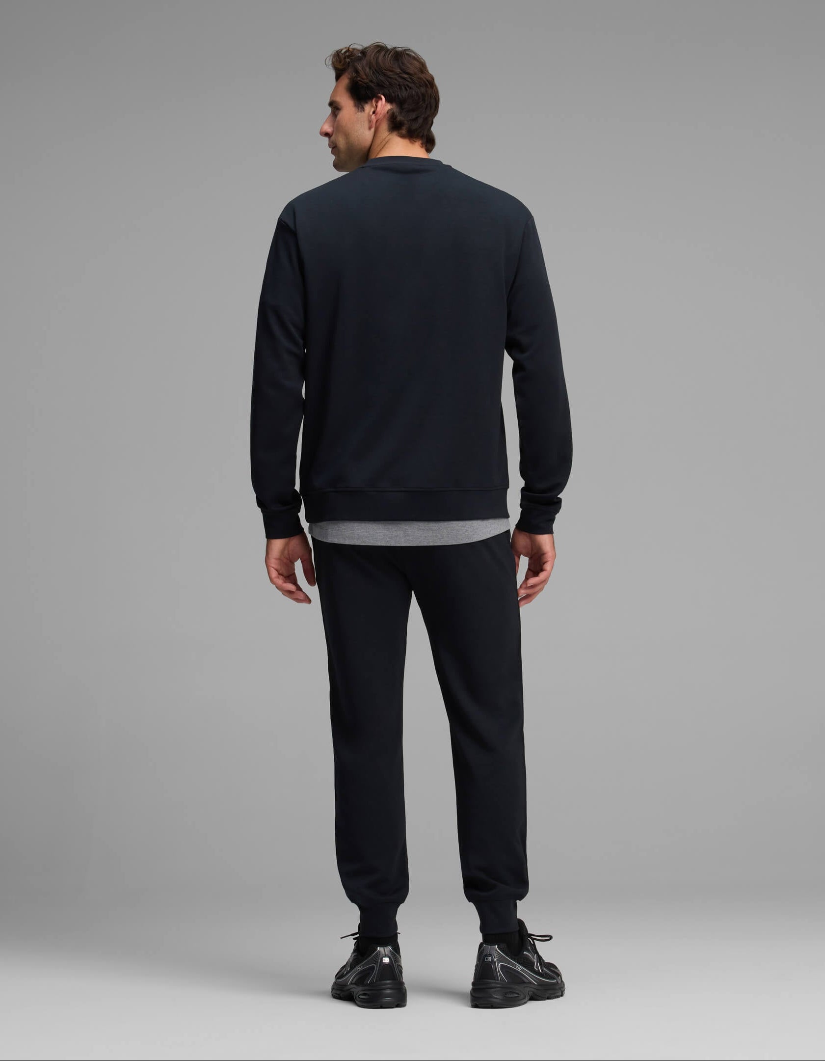 A man is standing with his back to the camera, wearing the Cozy Earth Men's Versa Crewneck & Jogger Set in black, paired with black sneakers and a gray shirt peeking out underneath. The background is plain and light gray.