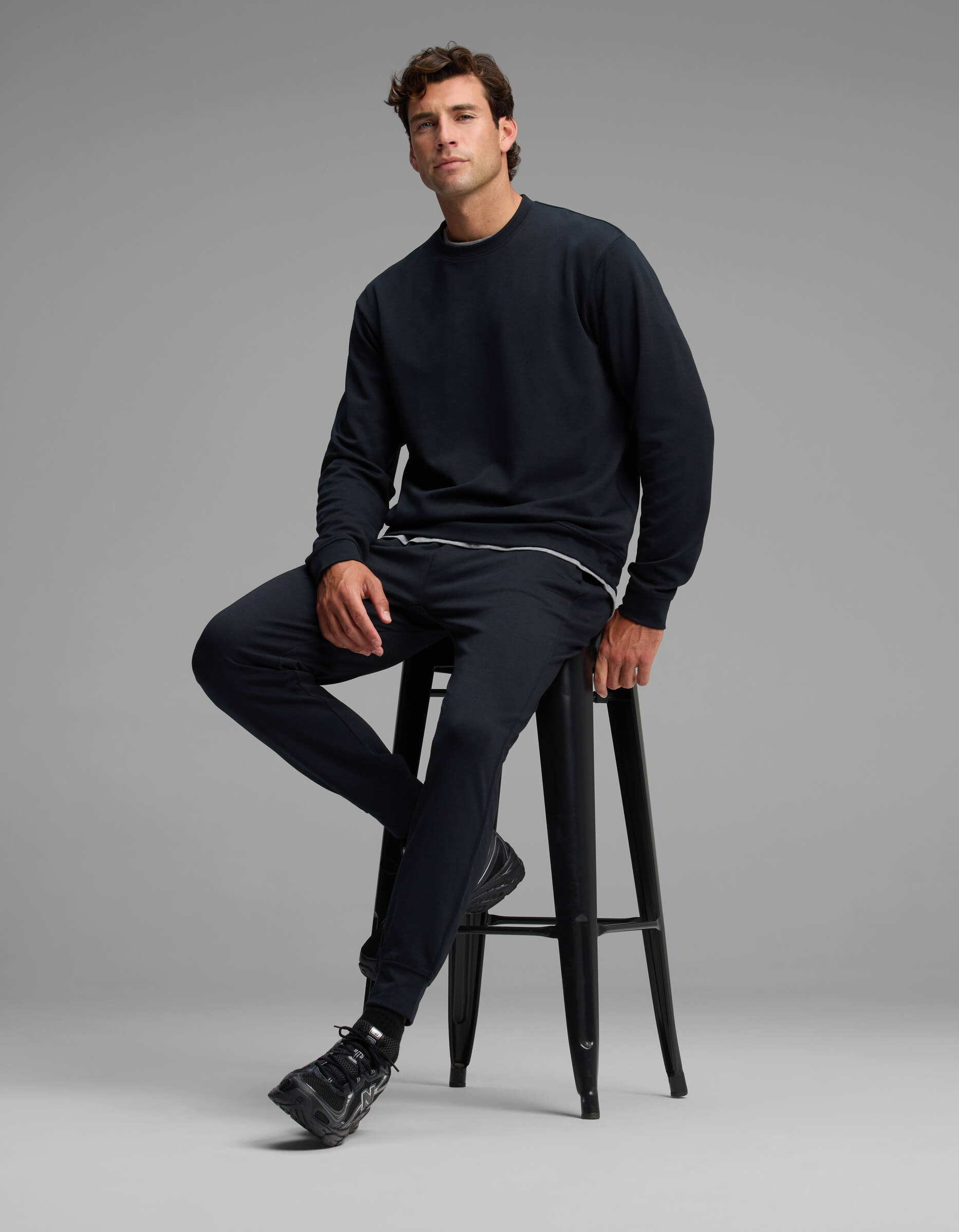 A man dressed in the Cozy Earth Men's Versa Crewneck & Jogger Set sits on a tall black stool against a plain gray background.