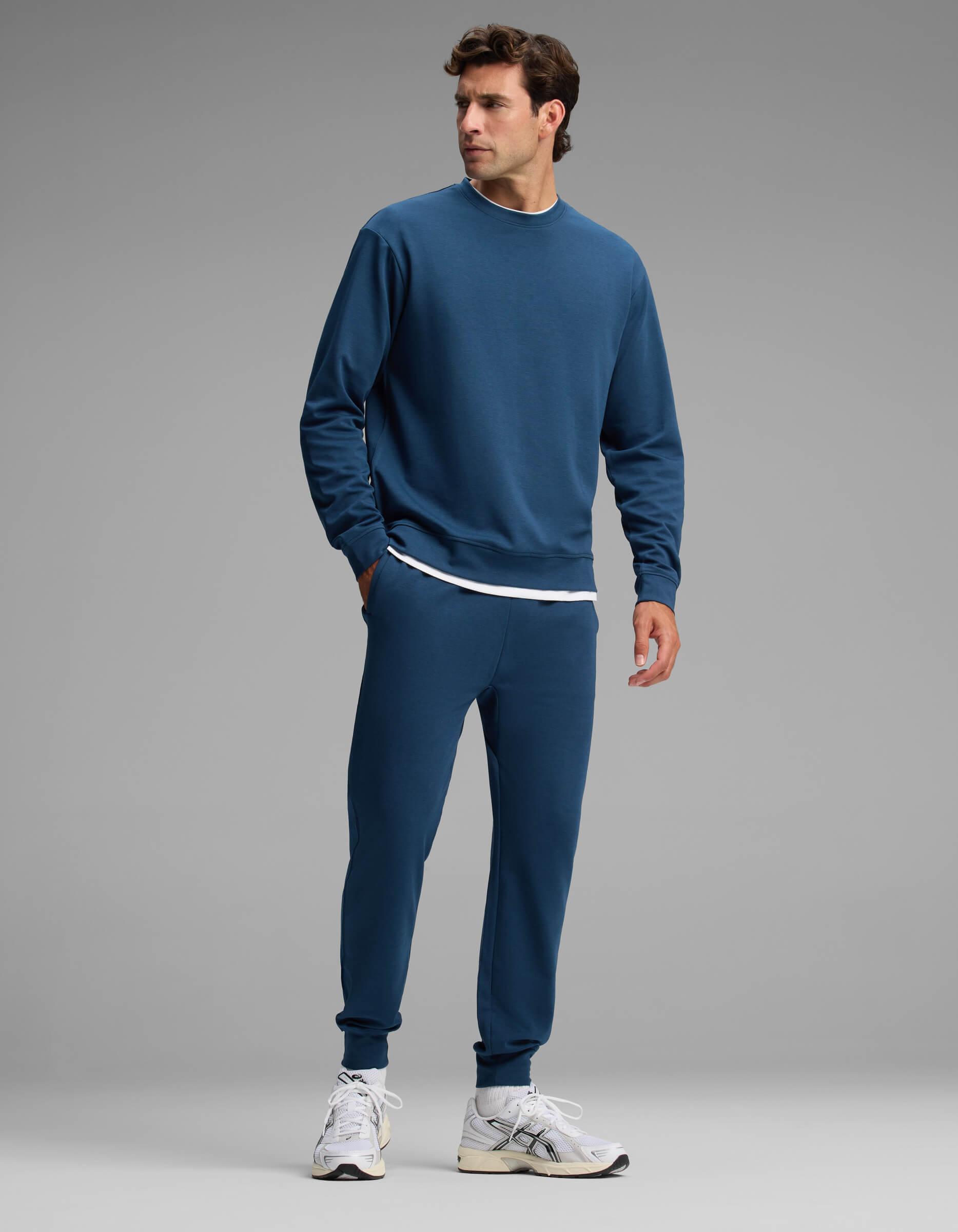 A man wearing the Cozy Earth Men's Versa Crewneck & Jogger Set in blue, paired with a white undershirt and white sneakers, stands against a plain gray background, looking to his right.
