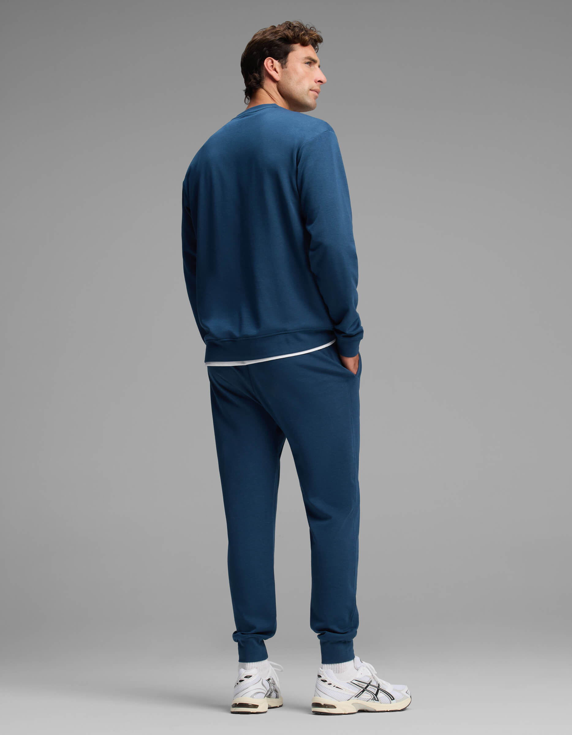 A man models the Cozy Earth Men's Versa Crewneck & Jogger Set in blue with white sneakers against a plain gray background. He stands facing away, hands in his jogger pockets, looking to his left.