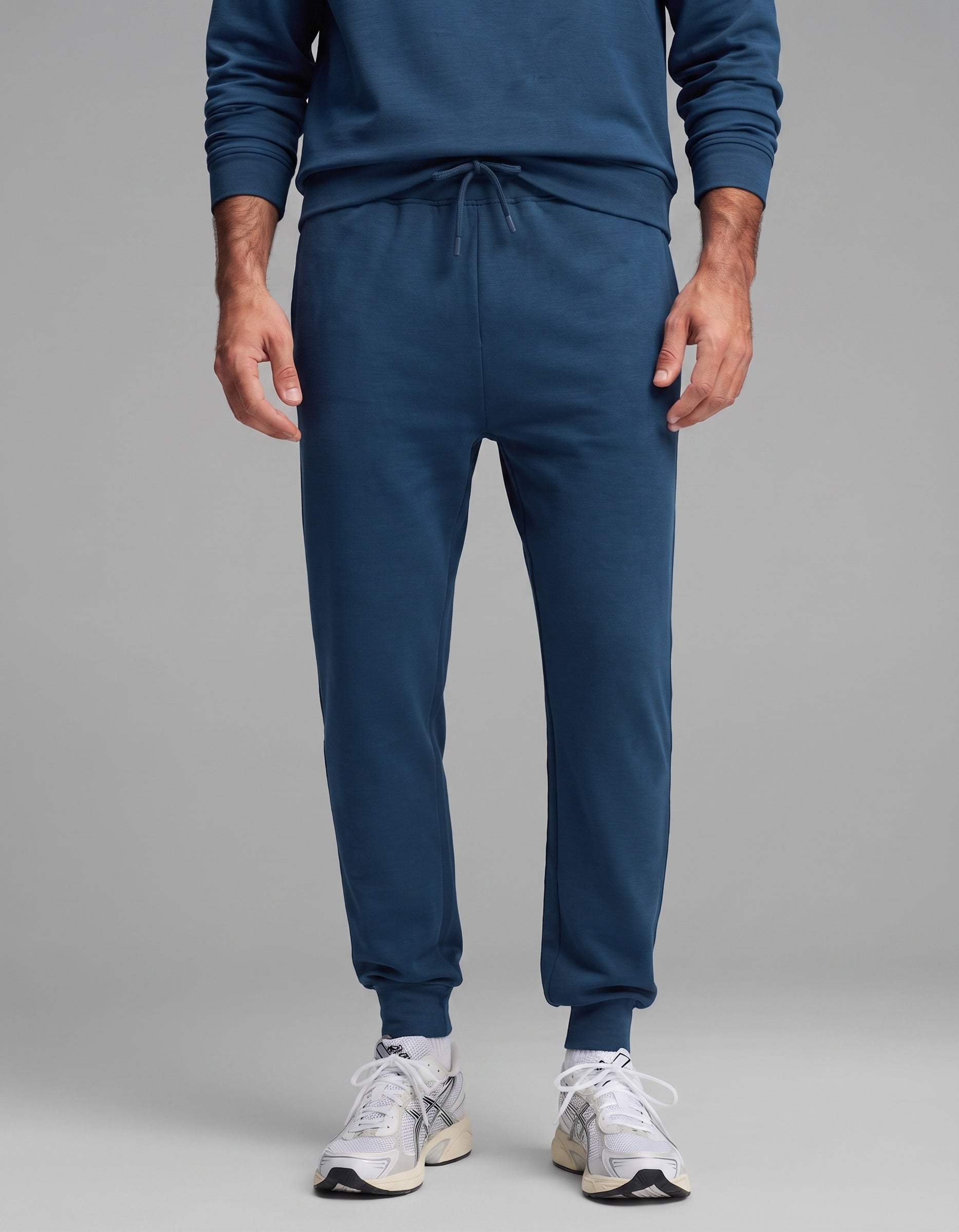 A person models the Cozy Earth Men's Versa Crewneck & Jogger Set in blue, paired with white athletic sneakers, standing against a plain gray background. The image is cropped at the chest.