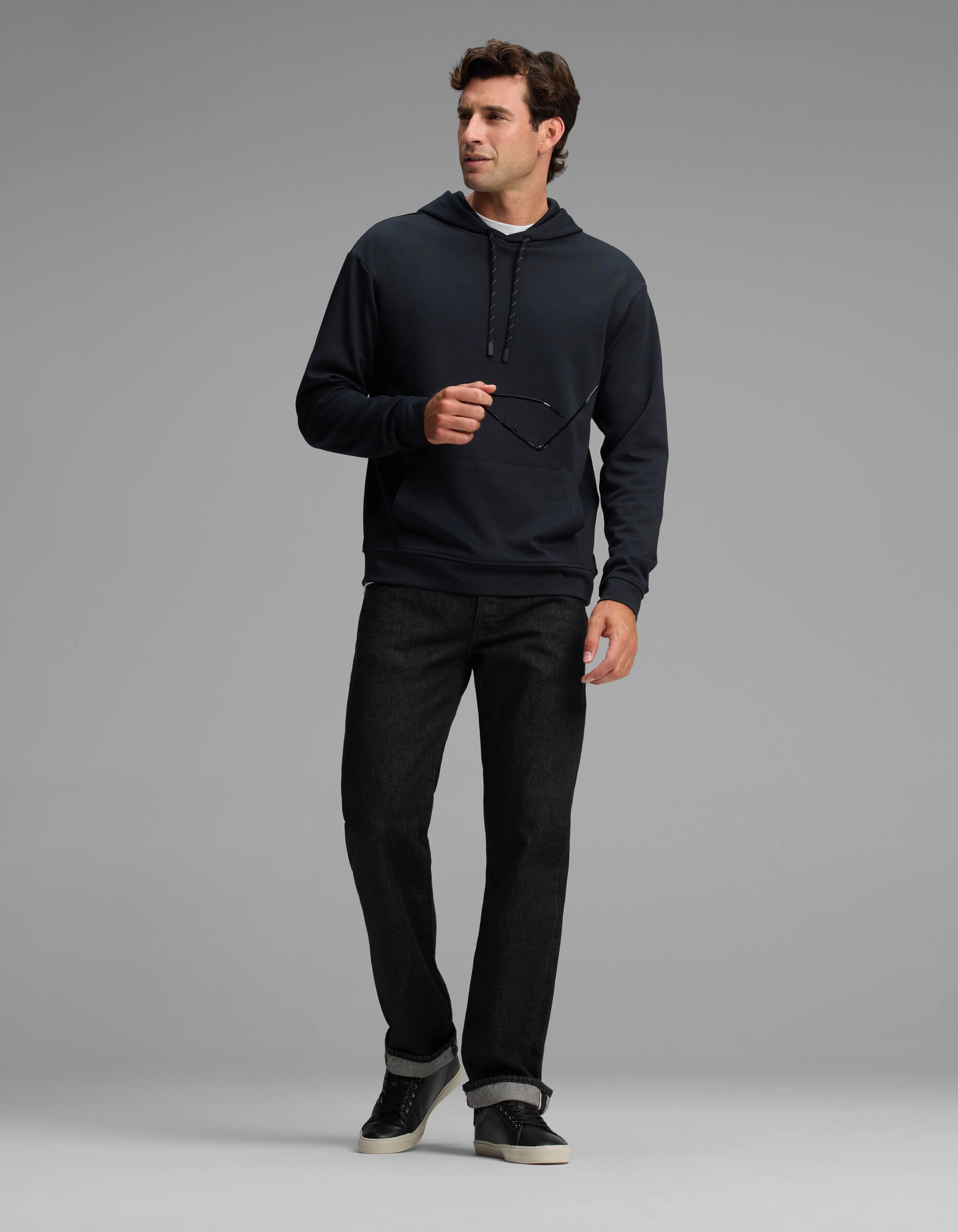 A man in the Cozy Earth Men's Versa Hoodie and black jeans stands against a plain gray background, holding eyeglasses in one hand and looking to the side.