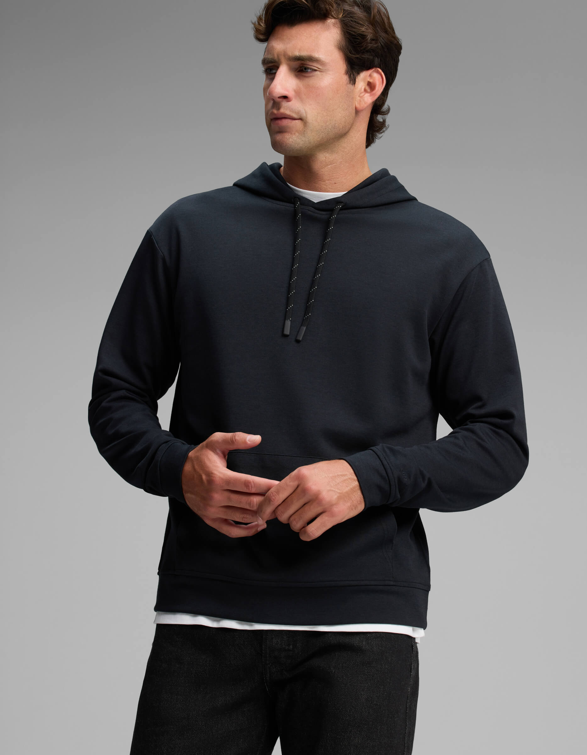 A man wearing the Cozy Earth Men's Versa Hoodie and dark jeans stands against a plain gray background, looking to his left with his hands loosely clasped in front of him.
