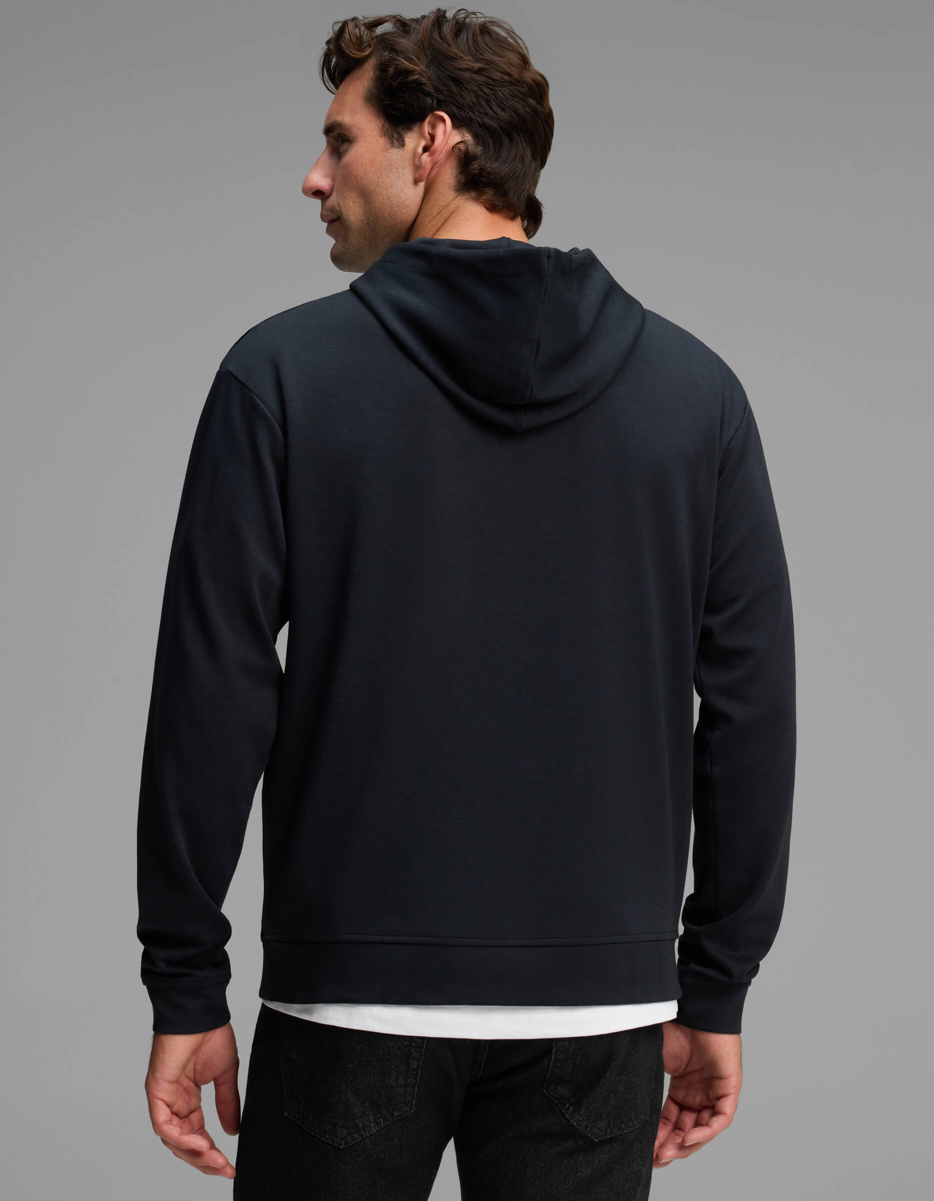 A man stands facing away against a plain gray background, wearing the Cozy Earth Men's Versa Hoodie in black over a white shirt and paired with black jeans.