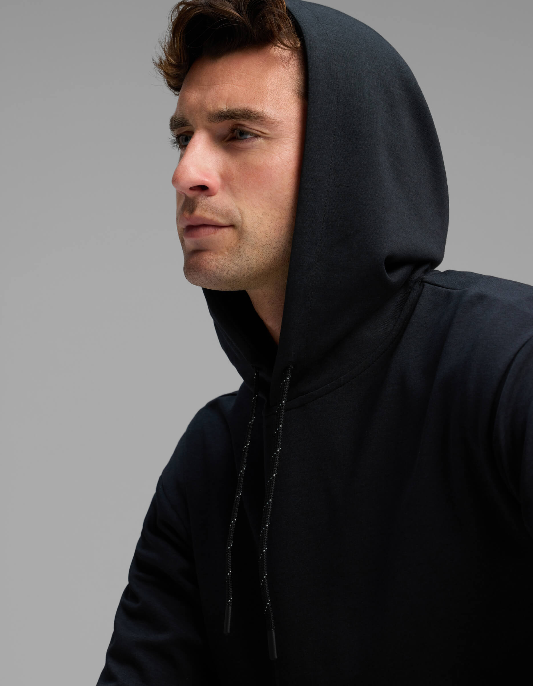 A man wearing the Cozy Earth Men's Versa Hoodie in black looks off to the side against a plain gray background.