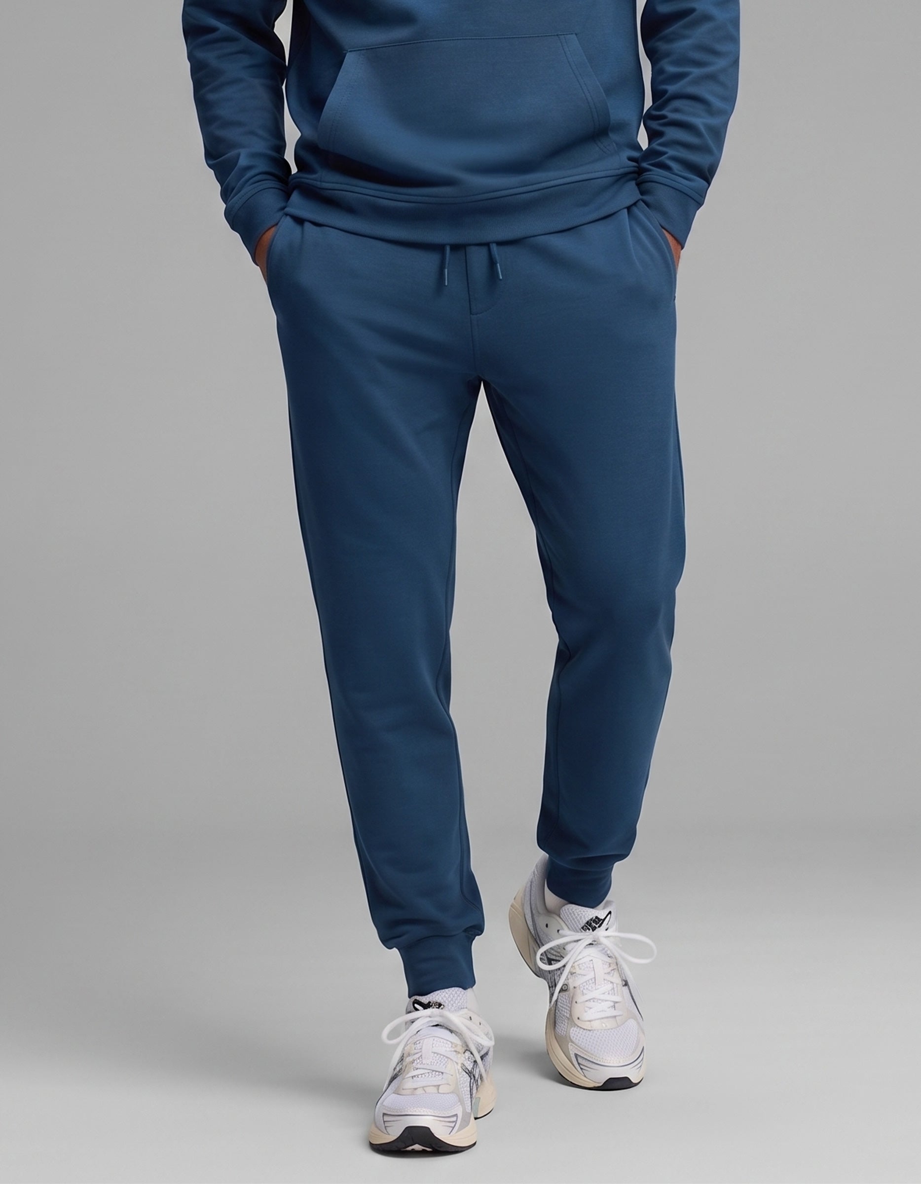Model is wearing the Cozy Earth Men's Versa Hoodie & Jogger Set in blue, paired with white athletic sneakers, standing with hands in pockets against a plain gray background.