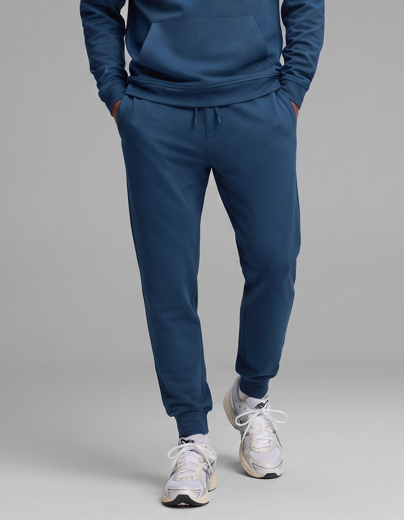 Model is wearing the Cozy Earth Men's Versa Hoodie & Jogger Set in blue, paired with white athletic sneakers, standing with hands in pockets against a plain gray background.