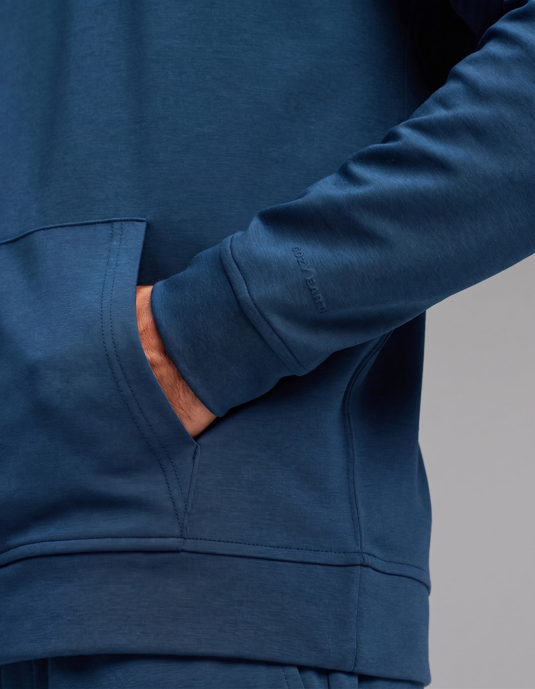 A person wearing the Cozy Earth Men's Versa Hoodie & Jogger Set in blue has their right hand in the front pocket, with the image highlighting the sleeve and pocket details.