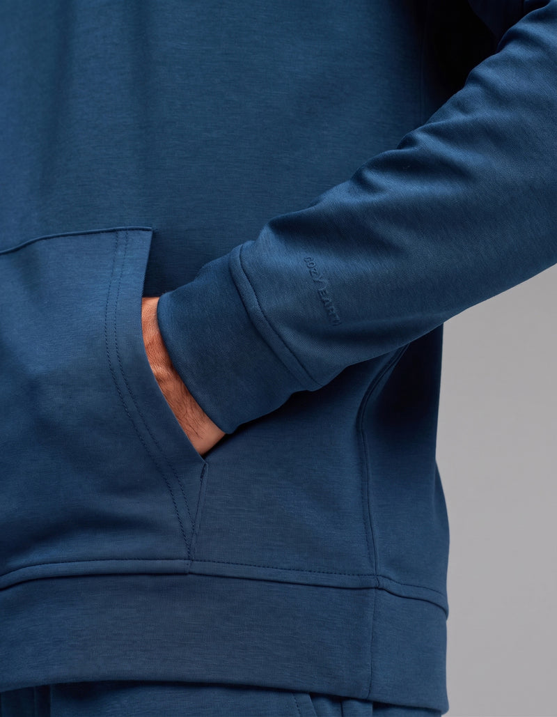 A person wearing the Cozy Earth Men's Versa Hoodie & Jogger Set in blue has their right hand in the front pocket, with the image highlighting the sleeve and pocket details.