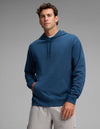 A man wearing the Cozy Earth Men's Versa Hoodie in blue and light gray shorts stands with one hand in his pocket against a plain gray background.|Color:Obsidian