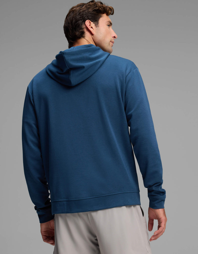 A man wearing the Cozy Earth Men's Versa Hoodie in blue and light gray shorts stands facing away, glancing over his left shoulder against a plain gray background.