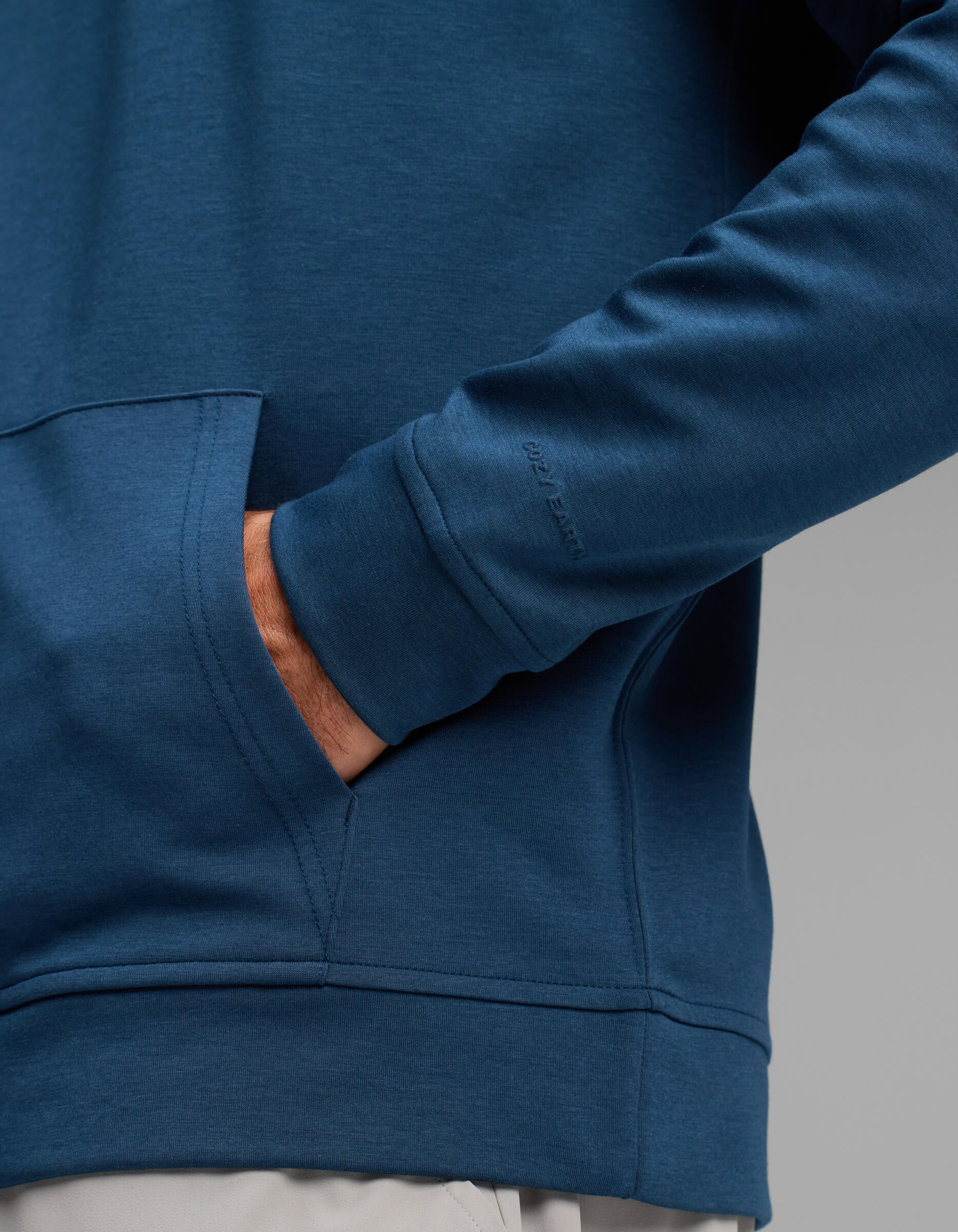 A man wearing the Cozy Earth Men's Versa Hoodie in blue, with one hand in the front pocket, stands against a plain gray background.