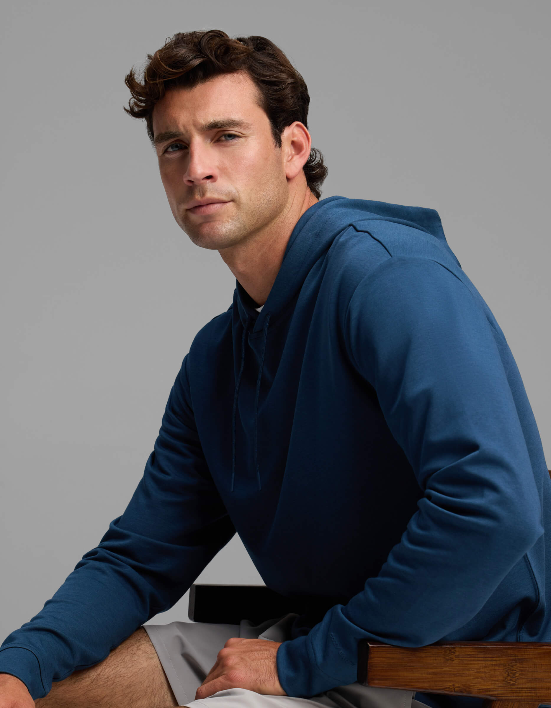 A man with short brown hair wears the Men's Versa Hoodie by Cozy Earth in blue and light gray shorts as he sits on a wooden chair, gazing slightly to the side with a neutral expression against a plain gray background.