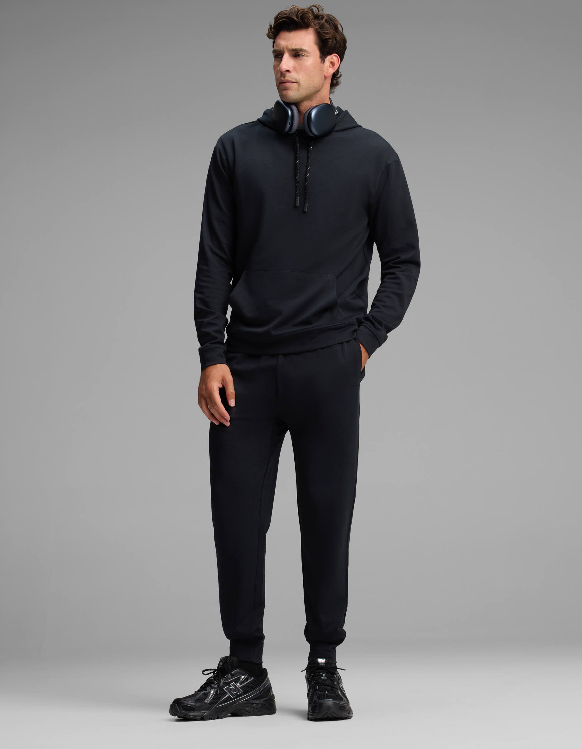 A man stands against a plain gray background, wearing the Cozy Earth Men's Versa Hoodie & Jogger Set in black, paired with black sneakers and over-ear headphones around his neck, looking slightly to the side.