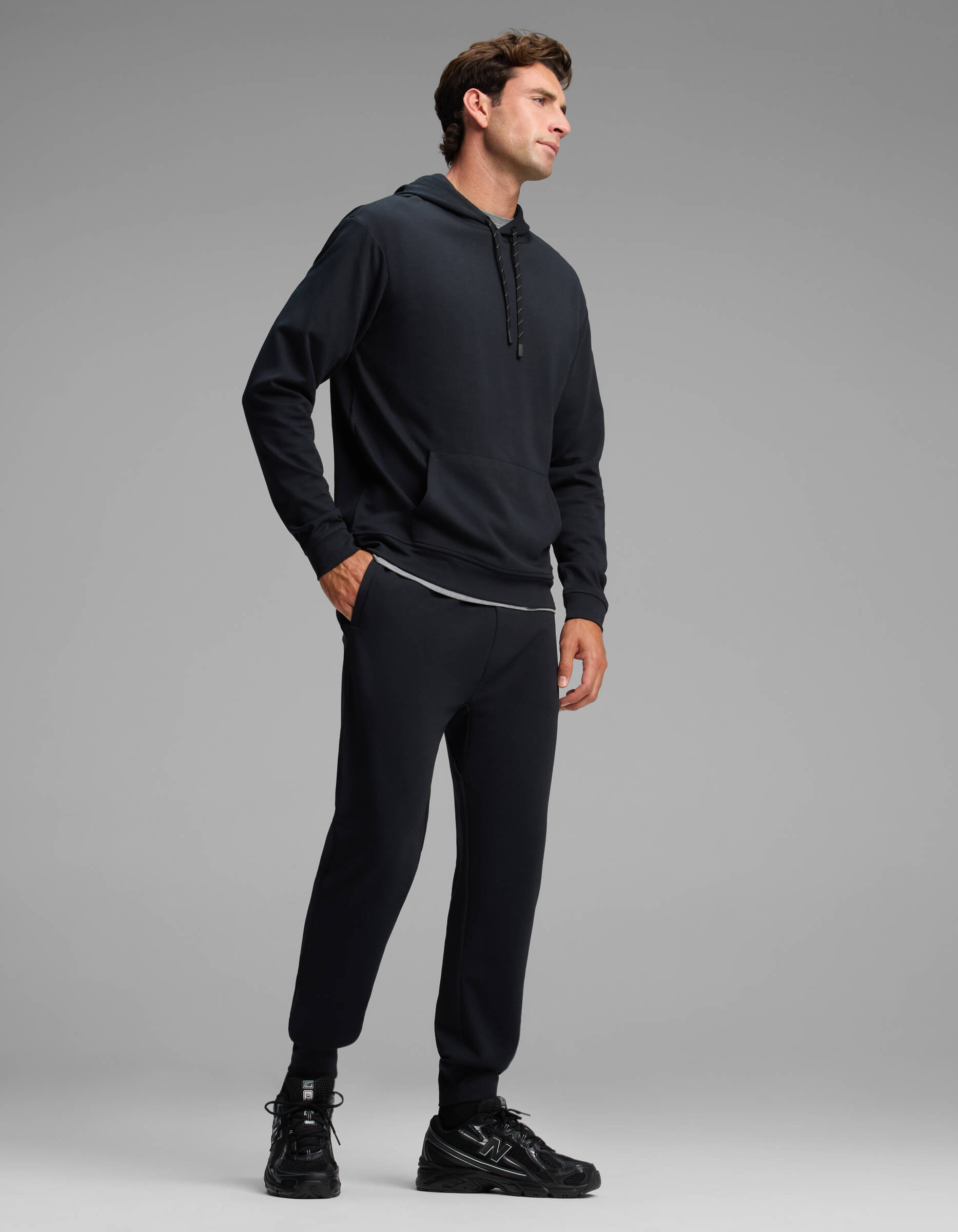 A man stands with his hands in his pockets, wearing the Cozy Earth Men's Versa Hoodie & Jogger Set in black, paired with black sneakers against a plain gray background.
