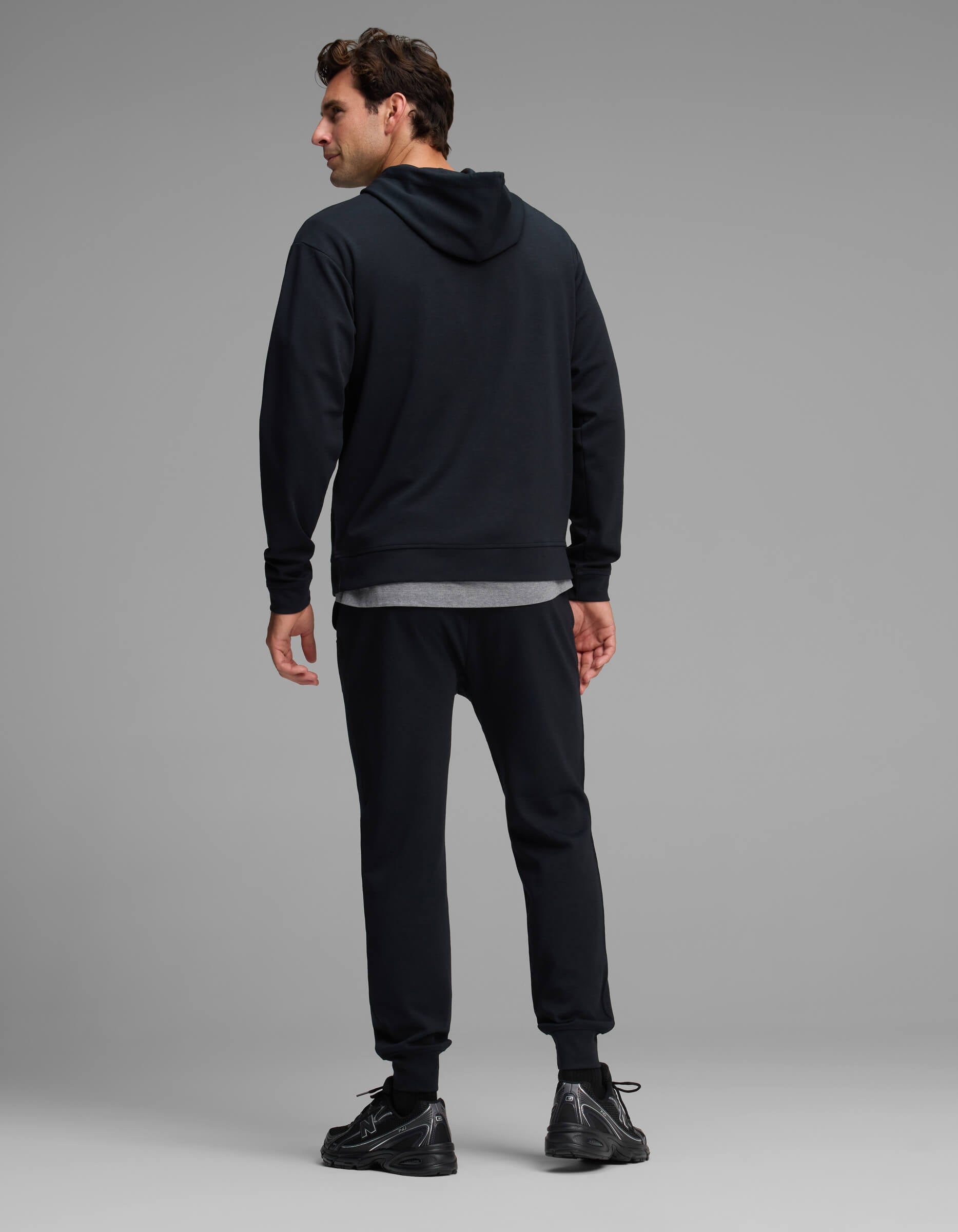 A man stands facing away, wearing the Cozy Earth Men's Versa Hoodie & Jogger Set in black. He is standing on a light gray floor with a plain gray background.