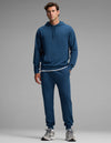 A man stands against a gray background wearing the Cozy Earth Men's Versa Hoodie & Jogger Set in blue, paired with a white T-shirt and gray sneakers. He looks forward with a neutral expression, one hand by his side and the other slightly bent.|Color:Obsidian
