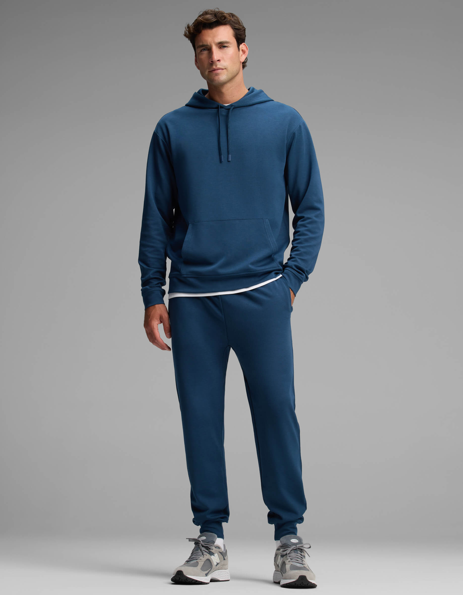 A man stands against a gray background wearing the Cozy Earth Men's Versa Hoodie & Jogger Set in blue, paired with a white T-shirt and gray sneakers. He looks forward with a neutral expression, one hand by his side and the other slightly bent.
