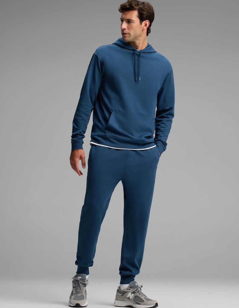 A man dressed in the Cozy Earth Men's Versa Hoodie & Jogger Set stands with one hand in his pocket, looking to the side. He completes the look with a white undershirt and gray sneakers.